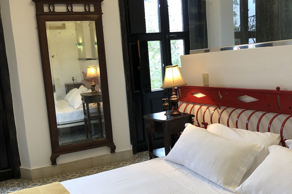 A hotel room with a large bed, a red headboard, a mirror, a nightstand with a lamp, and a window with a view of greenery.