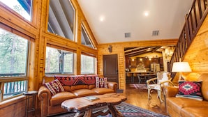 A cozy cabin interior with a leather sofa, wooden dining table, and a kitchen area visible through an open door. - Peaceful Family Mountain Lodge – Theater, Arcade, Hot Tub & Forest Views (Pine)
