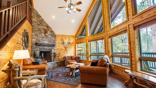 A cozy cabin interior with a stone fireplace, leather furniture, and large windows offering a view of the forest. - Peaceful Family Mountain Lodge – Theater, Arcade, Hot Tub & Forest Views (Pine)