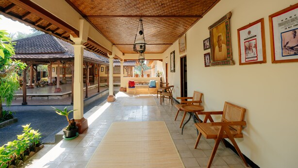 A covered outdoor seating area with wooden furniture, a tiled floor, and a view of a traditional building in the background. - BAKUNGAN diJero Homestay (Mengwi)