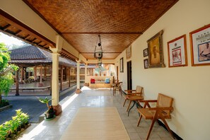 A covered outdoor seating area with wooden furniture, a tiled floor, and a view of a traditional building in the background. - BAKUNGAN diJero Homestay (Mengwi)