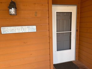 Une façade en bois avec une porte blanche, une pancarte d’accueil et une lanterne.
