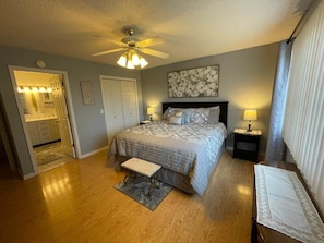 A bedroom with a bed, bedside tables, a bench, a ceiling fan, and a door leading to a bathroom.
