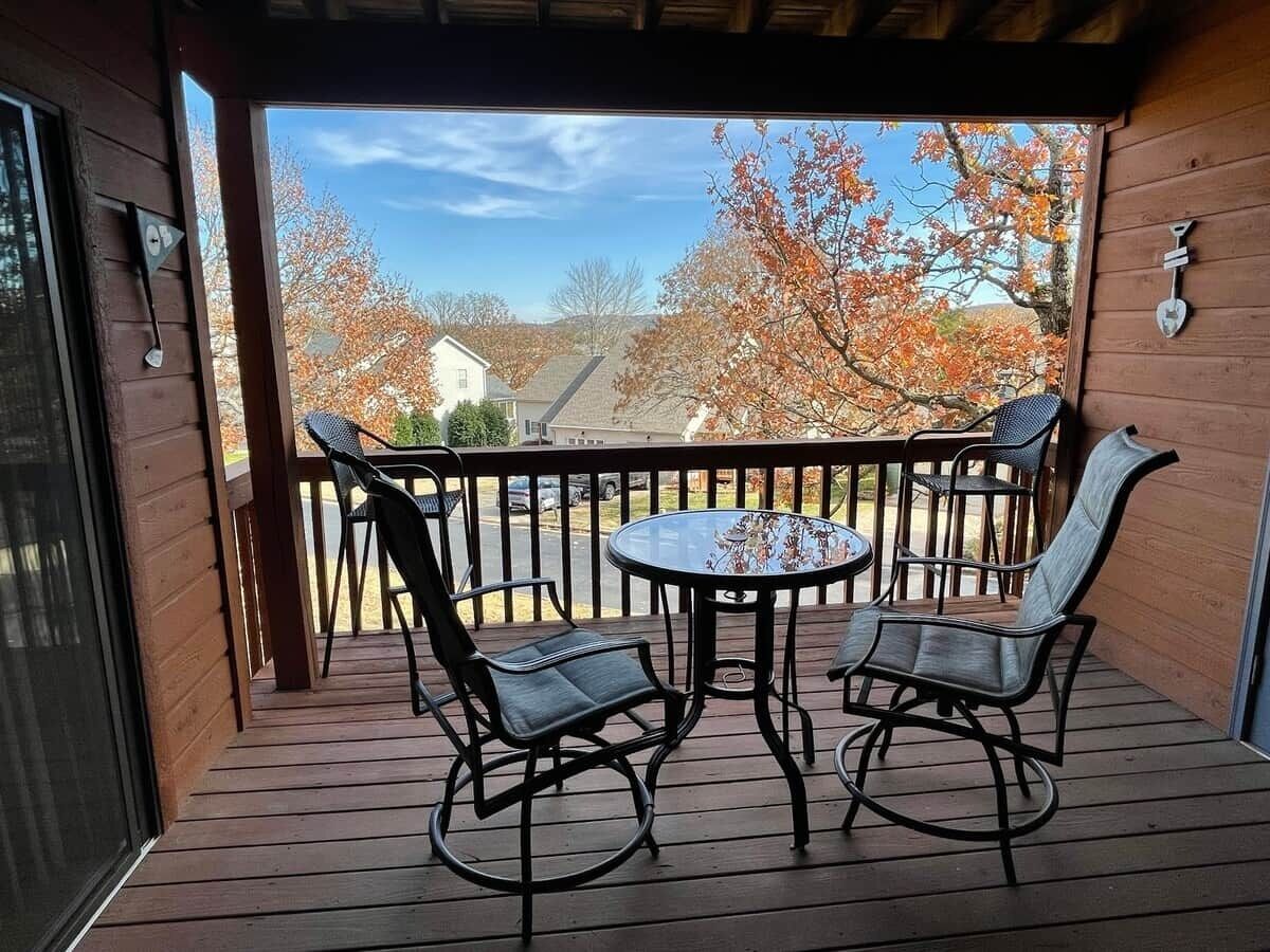 A wooden deck with a round table and two chairs, overlooking a residential area with autumn trees.