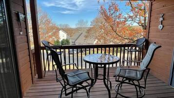 A wooden deck with a round table and two chairs, overlooking a residential area with autumn trees.