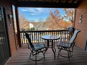 A wooden deck with a round table and two chairs, overlooking a residential area with autumn trees.