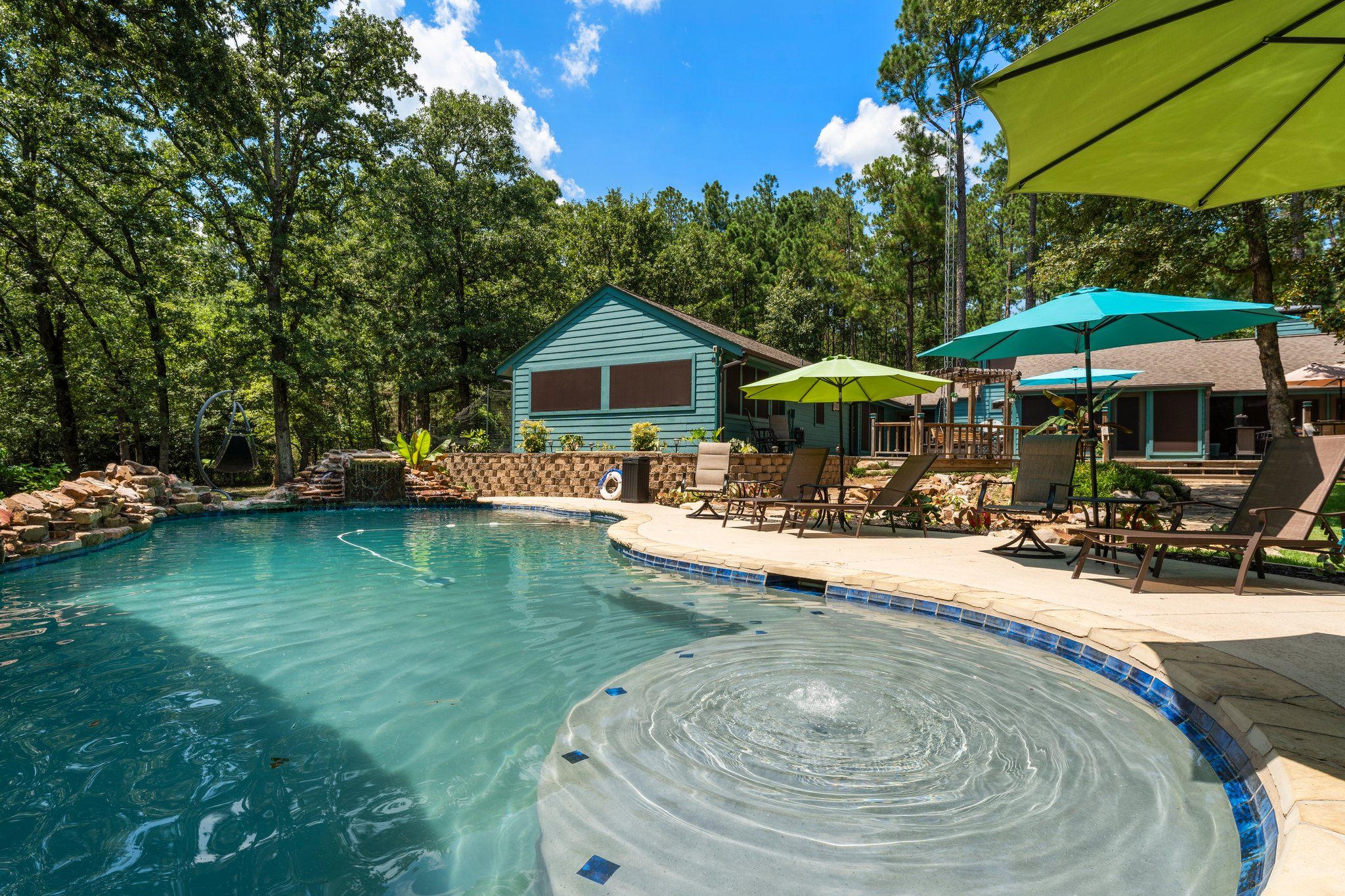 Ein Swimmingpool mit Steinrand, umgeben von Liegestühlen und Sonnenschirmen, mit einer Holzterrasse und einem Haus im Hintergrund.