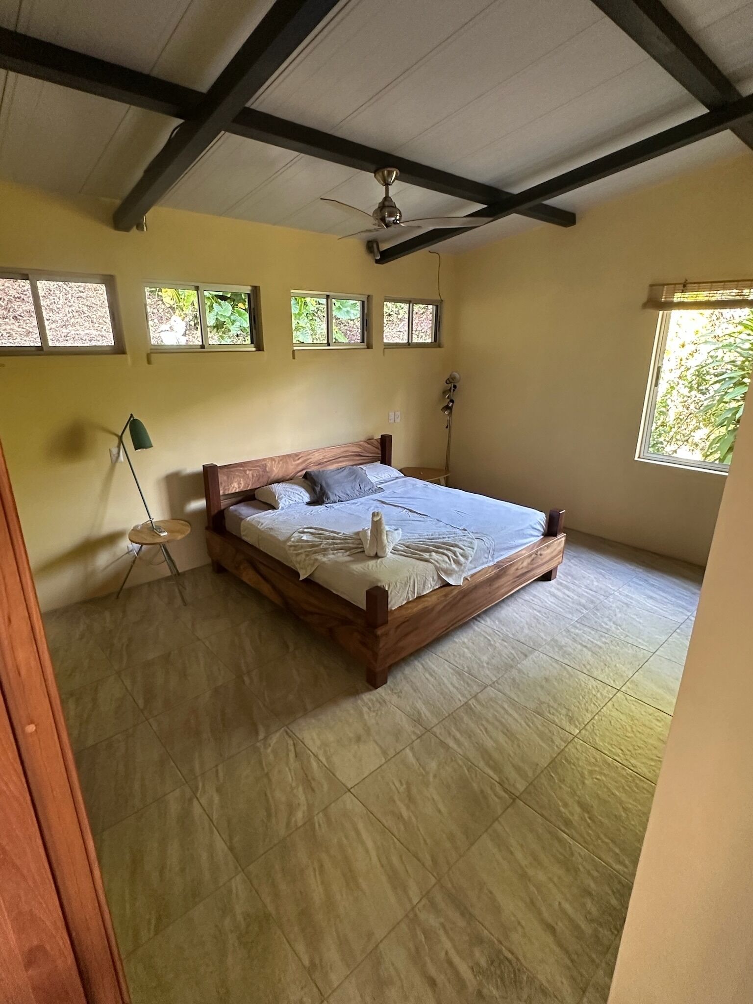 A bedroom with a wooden bed, a ceiling fan, and a window with a view of greenery.