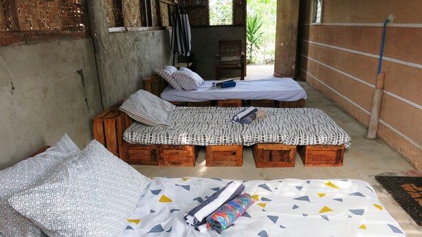 A rustic room with two wooden beds, one with a patterned bedspread and pillows, and another with a plain bedspread. There are wooden crates under the beds, and a large bed with a colorful geometric bedspread and a rolled-up blanket.