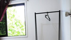A room with a standing fan, a window with curtains, a tiled floor, and a small shelf with folded clothes.