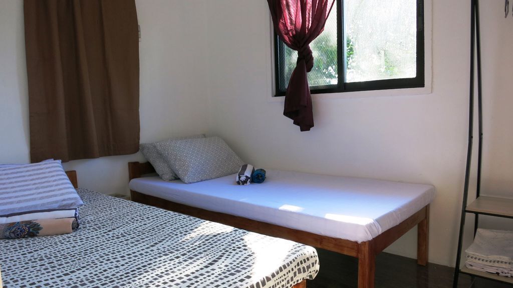 Ein kleiner, schlichter Raum mit einem Bett, einem Fenster mit Vorhängen und einem Regal mit gefalteten Handtüchern.