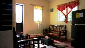 A compact living space with a wooden bench, a small table, a refrigerator, and a window with curtains.