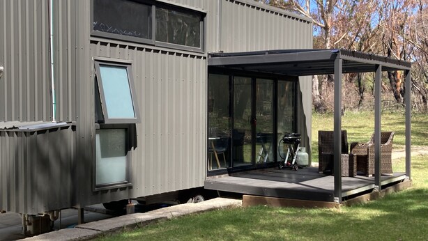 Un bâtiment moderne et compact, doté d’un espace extérieur couvert avec des sièges, de grandes fenêtres et d’une bouteille de gaz visible.