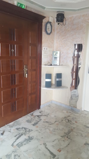 A room with a wooden door, marble flooring, a clock, electrical outlets, and two speakers on a shelf.