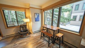 A room with a wooden floor, a rocking chair, a table with benches, and large windows offering a view of trees and buildings outside. - Mountain Retreat | Balcony, Hot Tub & Firepit (Keystone)