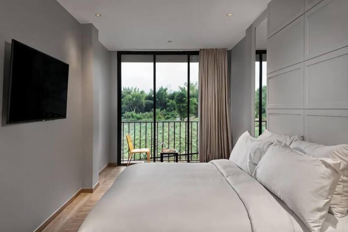 A bedroom with a large bed, a TV mounted on the wall, and a balcony with a view of greenery.