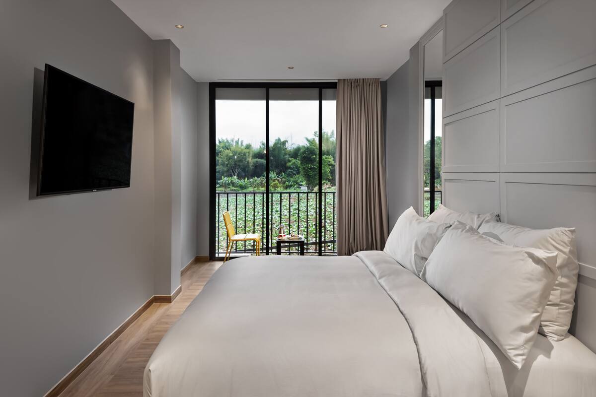 A modern bedroom with a large bed, a flat-screen TV, and a balcony with a view of greenery.