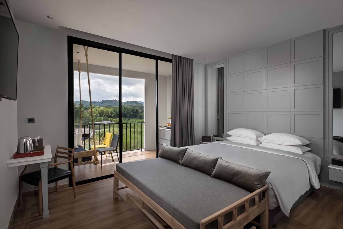 A modern hotel room with a large bed, a desk, and a balcony with a view of greenery.