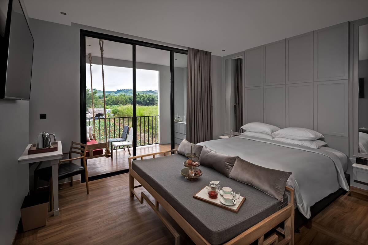 A modern bedroom with a large bed, a desk, and a balcony with a view of greenery.