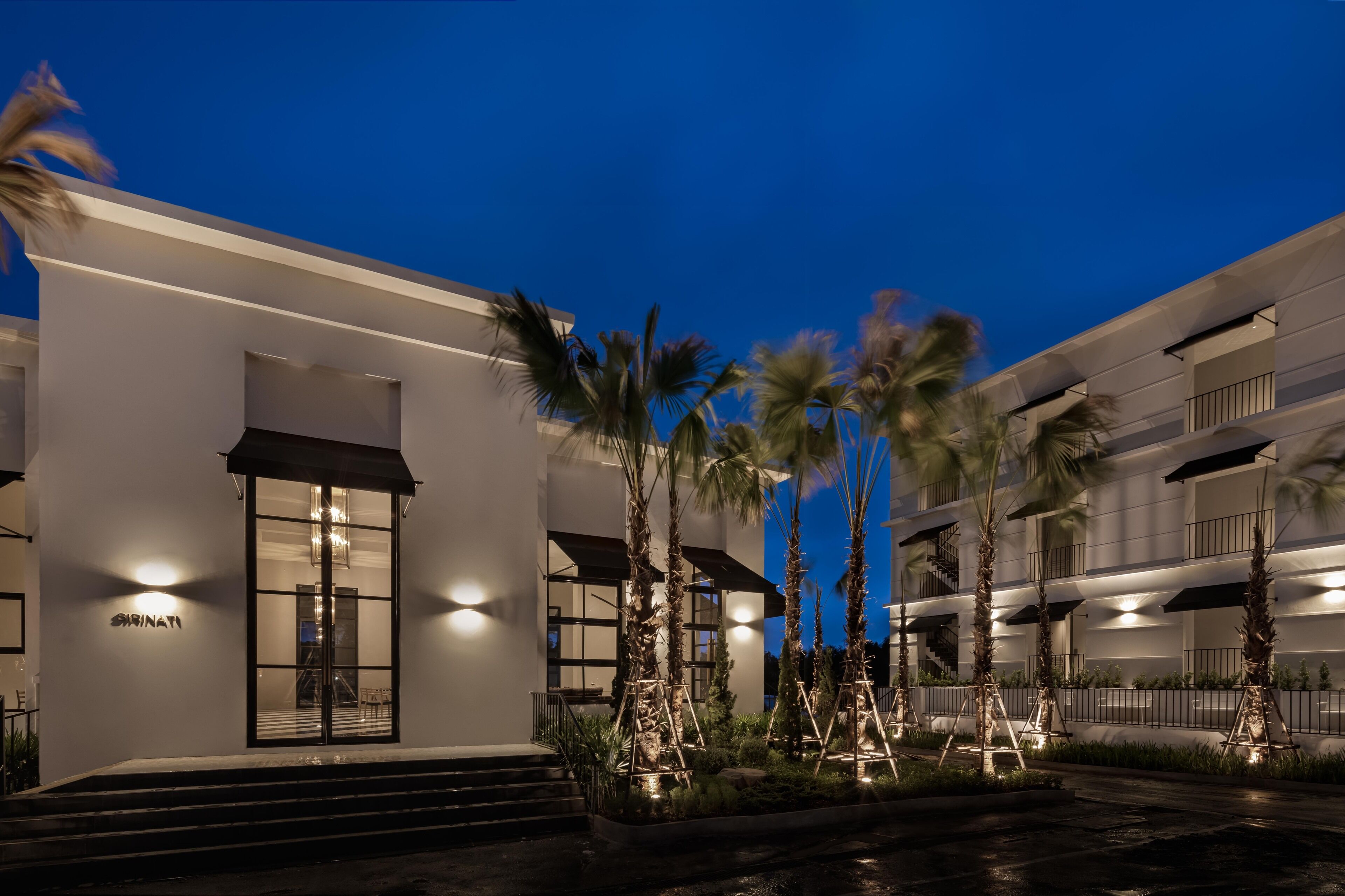 A modern hotel with a white facade, a glass entrance, and palm trees in the foreground.