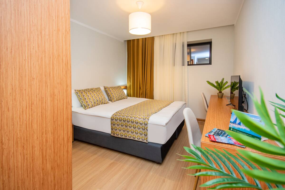 A hotel room with a bed, a desk with a computer, a mirror, and a window with curtains.