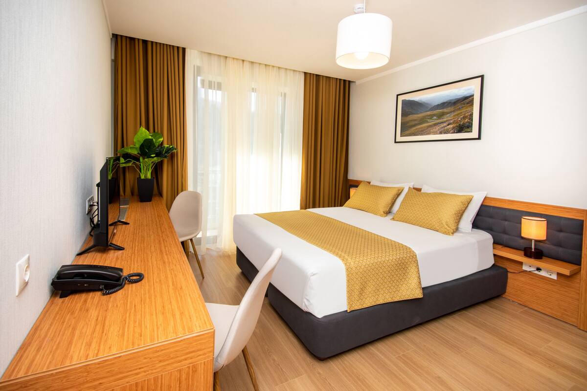 A hotel room with a bed, desk, chair, and a plant.