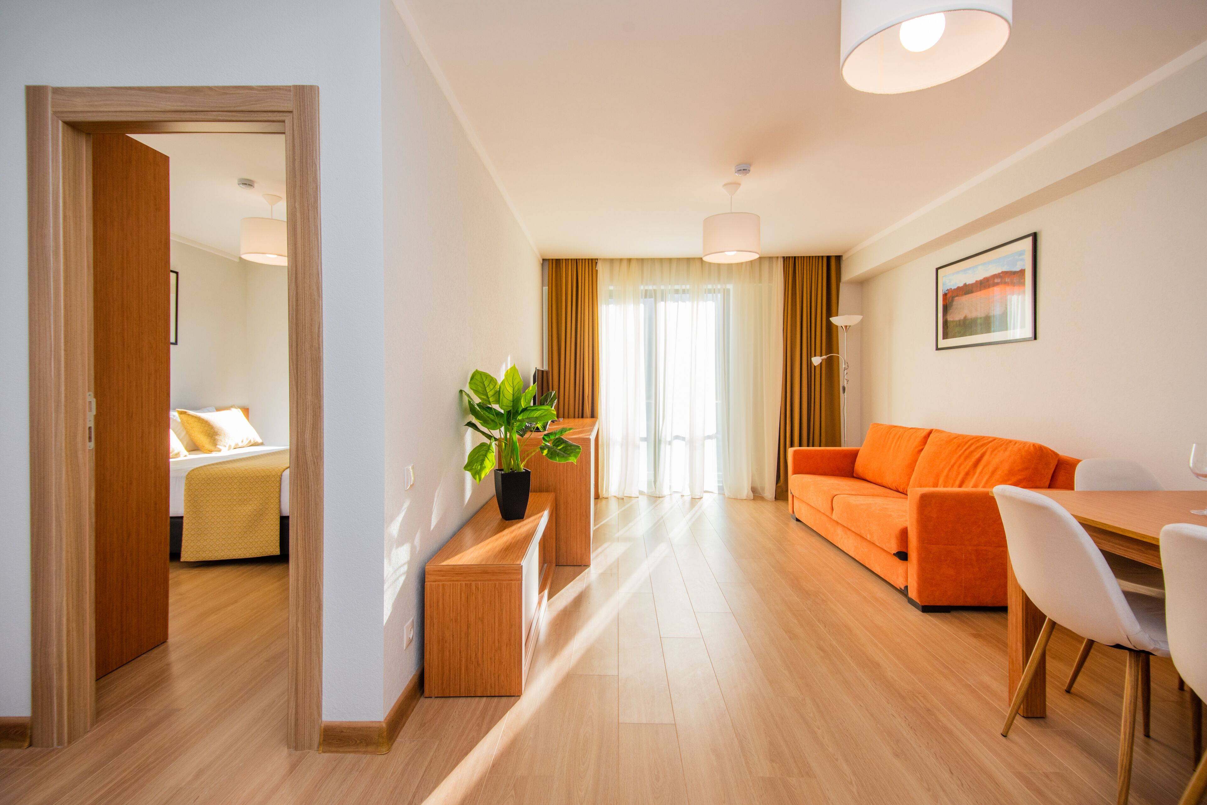 Una habitación de hotel moderna con suelo de madera, una cama con ropa de cama blanca, un banco de madera con una planta, un sofá naranja, una mesa de comedor con sillas blancas y un ventanal con cortinas.