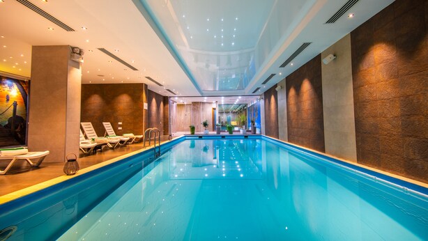 A large indoor swimming pool with lounge chairs and a view of a cityscape through a glass ceiling.