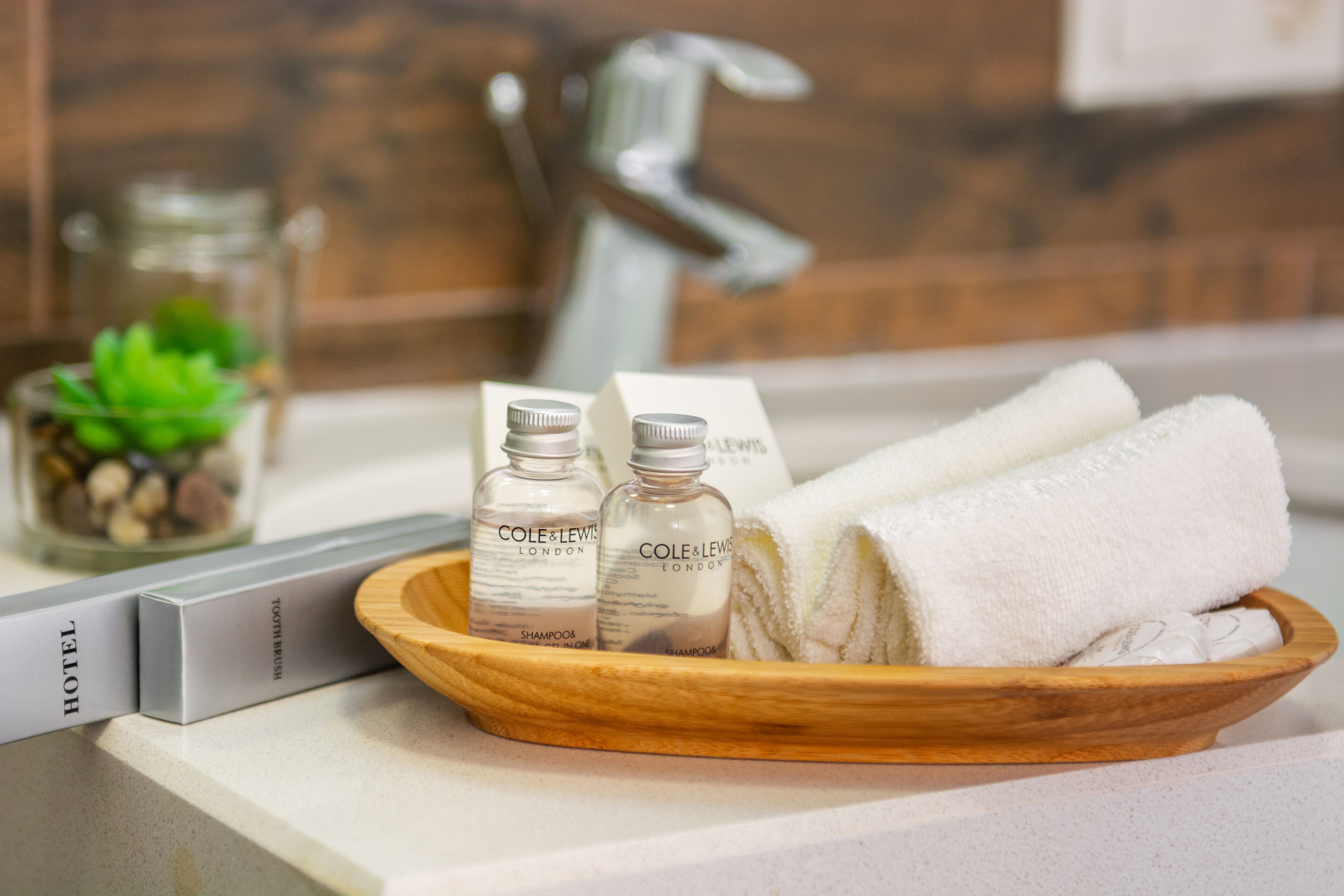 Een houten dienblad met flesjes shampoo, een opgevouwen handdoek en een boek met de titel 'HOTEL' op een aanrecht.