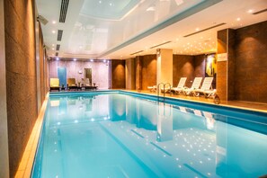 An indoor swimming pool with clear water, lounge chairs, and a reception area with a door.