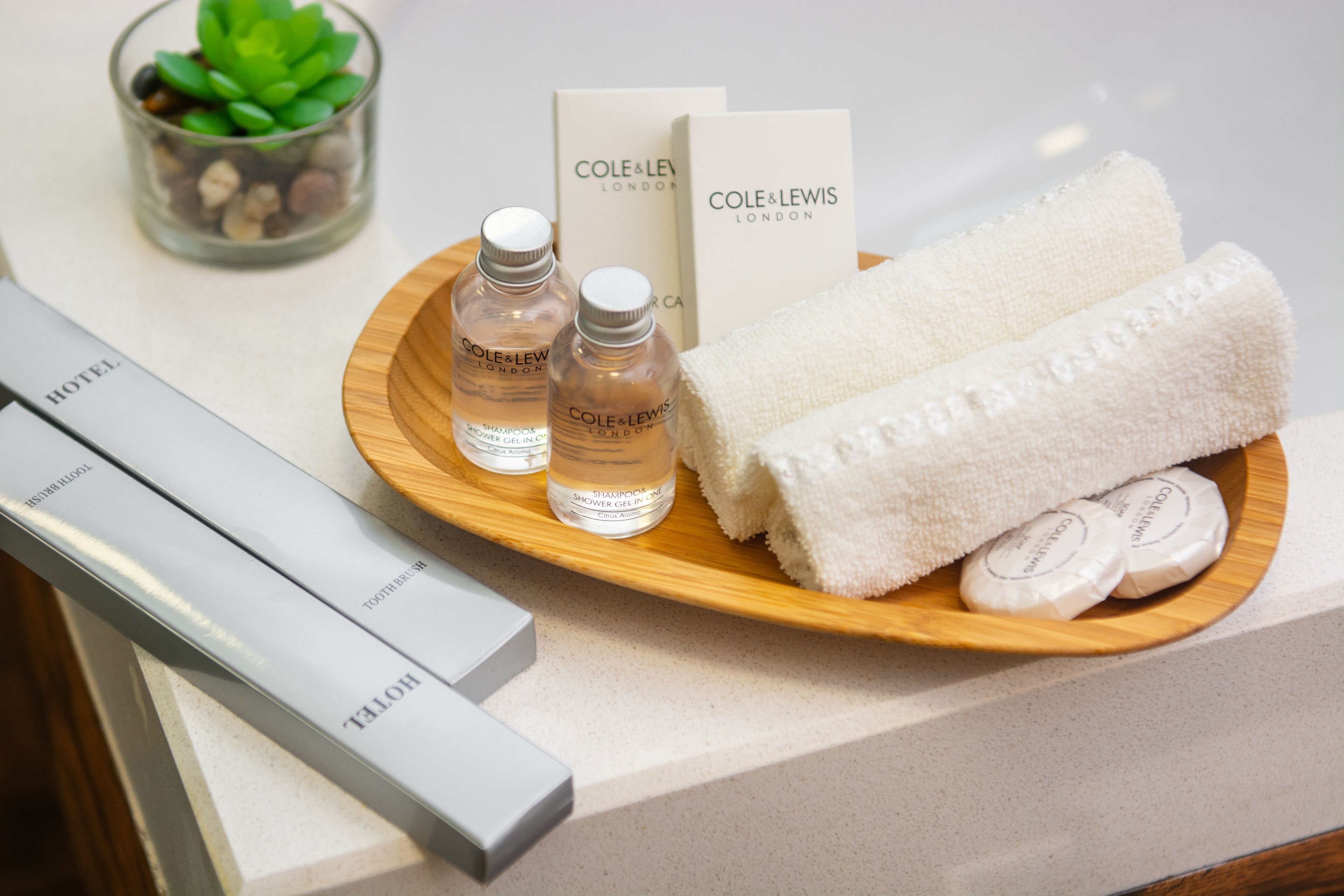 A hotel bathroom with toiletries on a wooden tray, including bottles of shampoo and conditioner, a towel, and a glass of water.