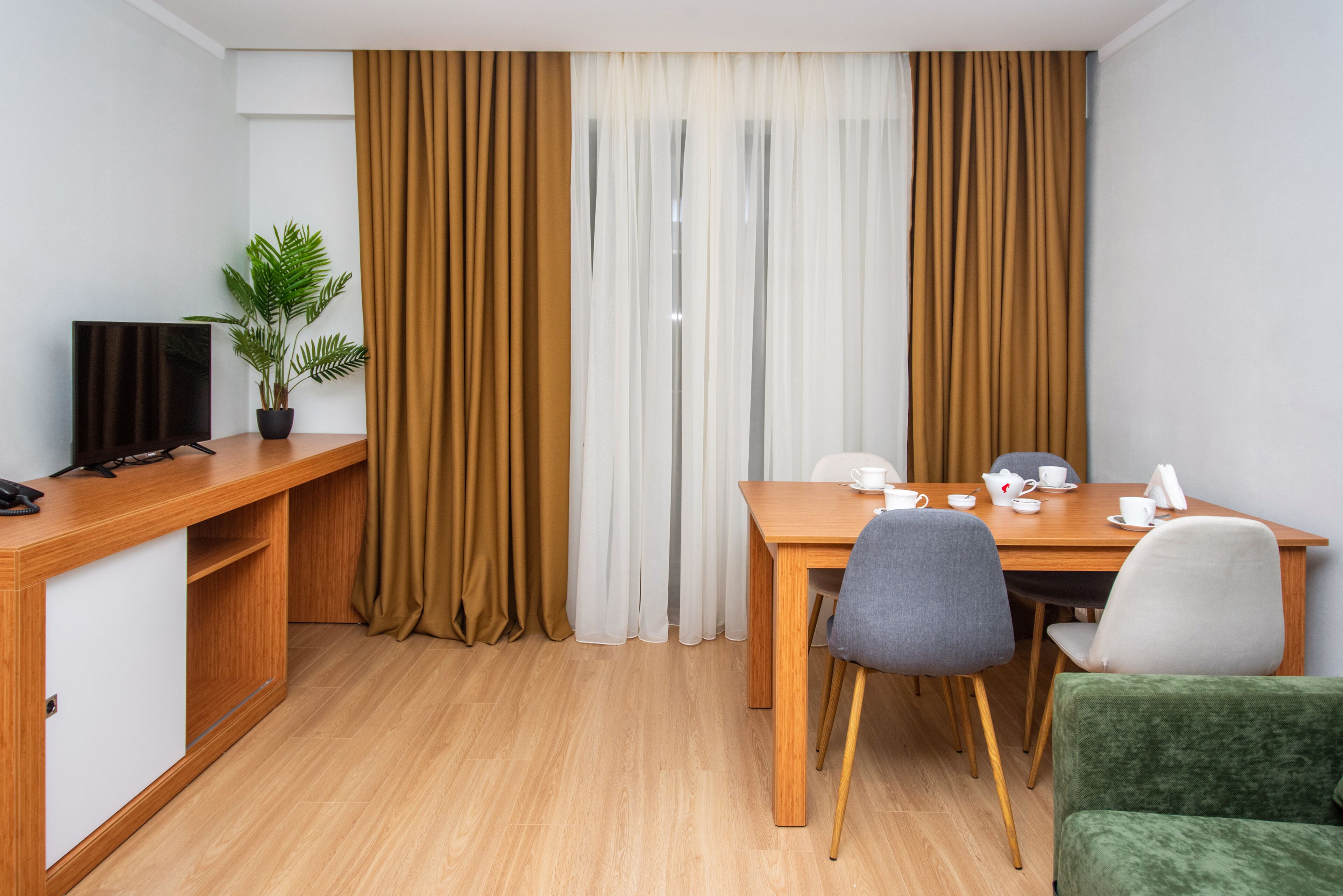 Une chambre d’hôtel avec un bureau en bois, une télévision, une plante en pot, une table à manger avec des chaises et une fenêtre avec des rideaux.