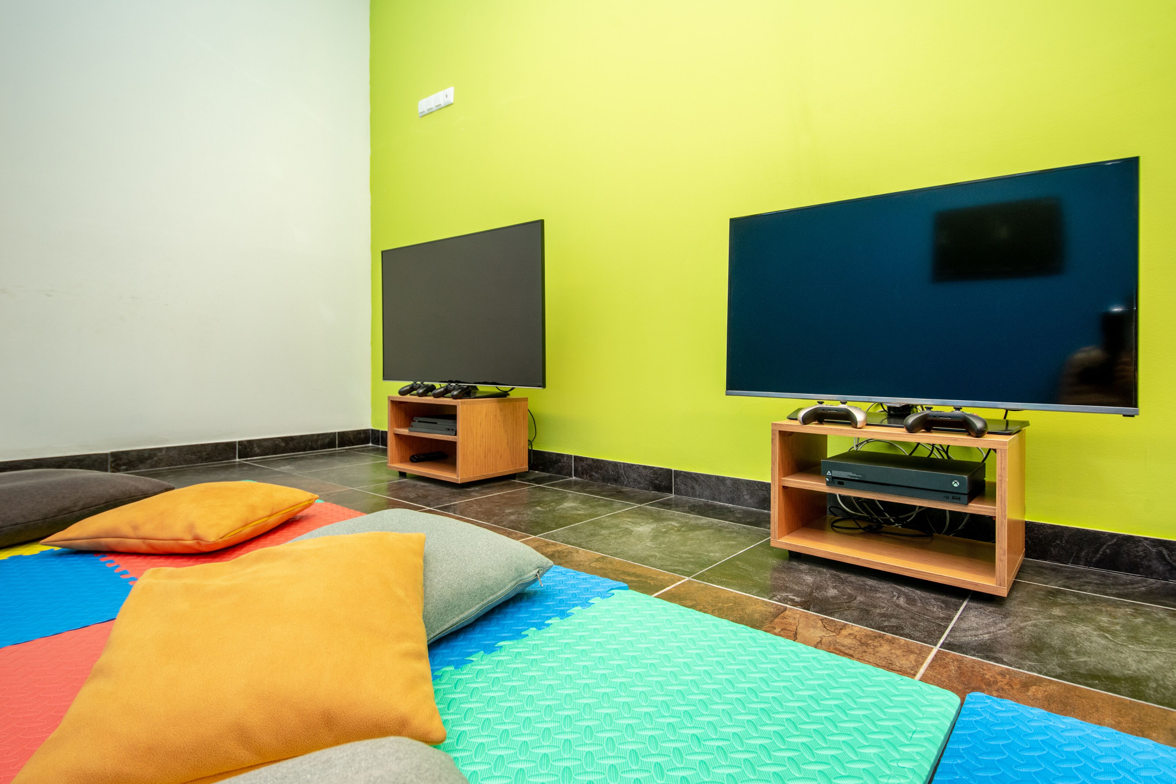 Una habitación con dos televisores sobre pedestales de madera, una alfombra de juego colorida y un pequeño estante con dispositivos electrónicos.