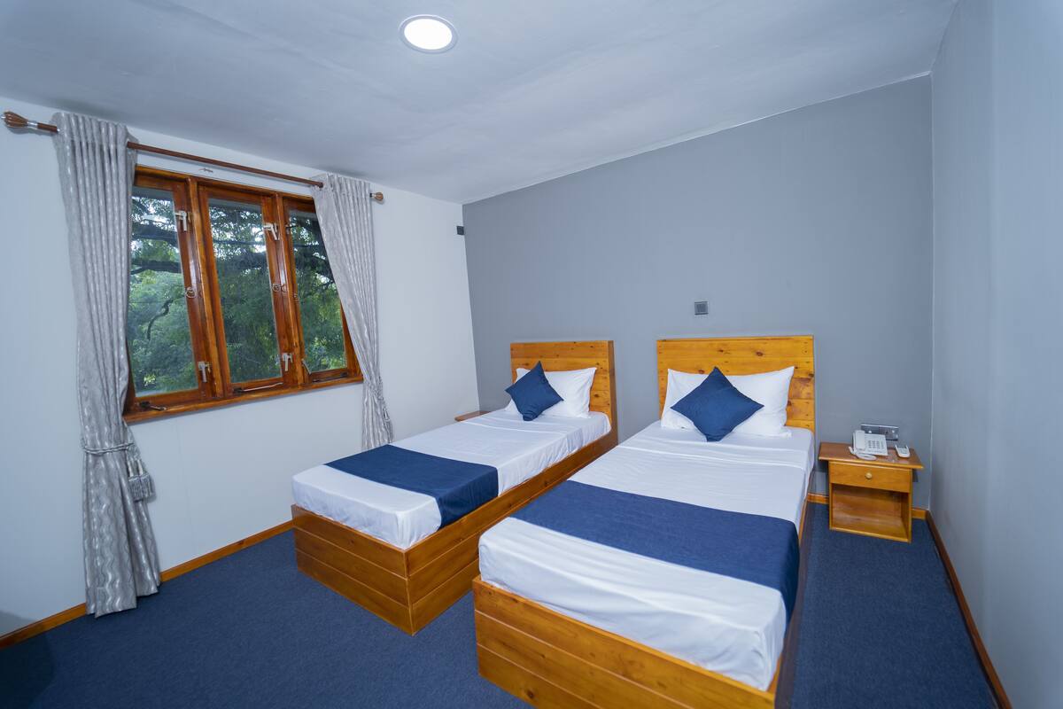 A room with two wooden beds, a window with curtains, a bedside table with a phone, and a carpeted floor.
