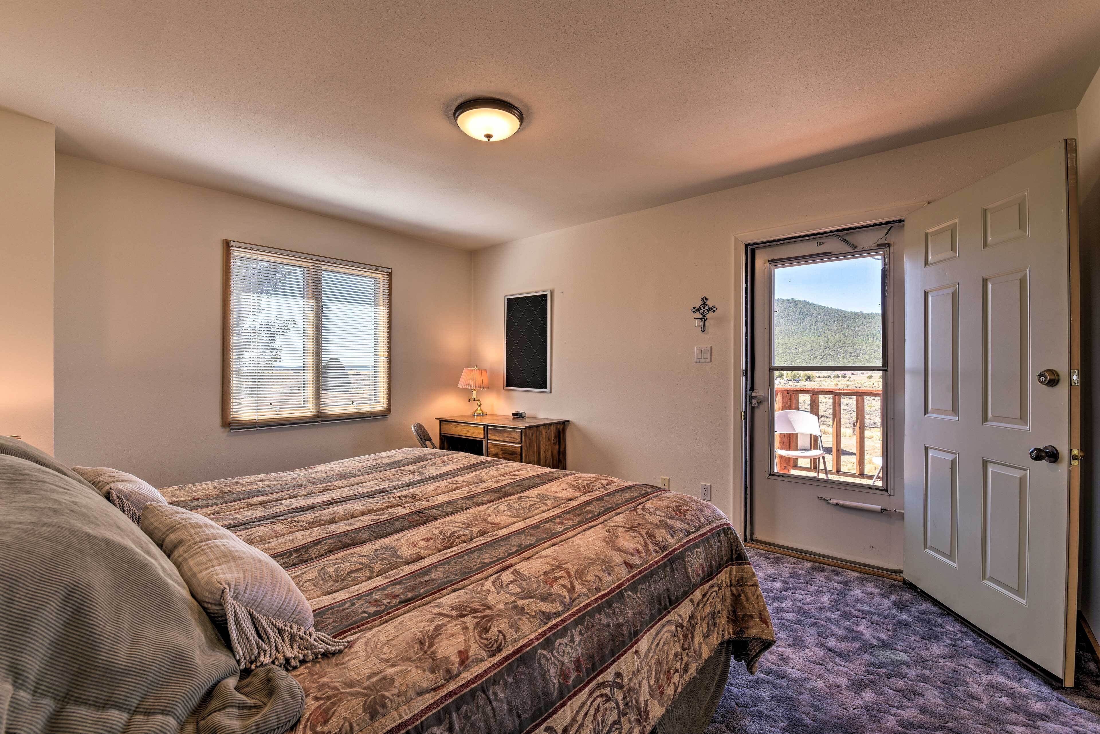 A bedroom with a large bed, a desk, a chair, a window with blinds, a door leading to an outdoor area, and a ceiling light.