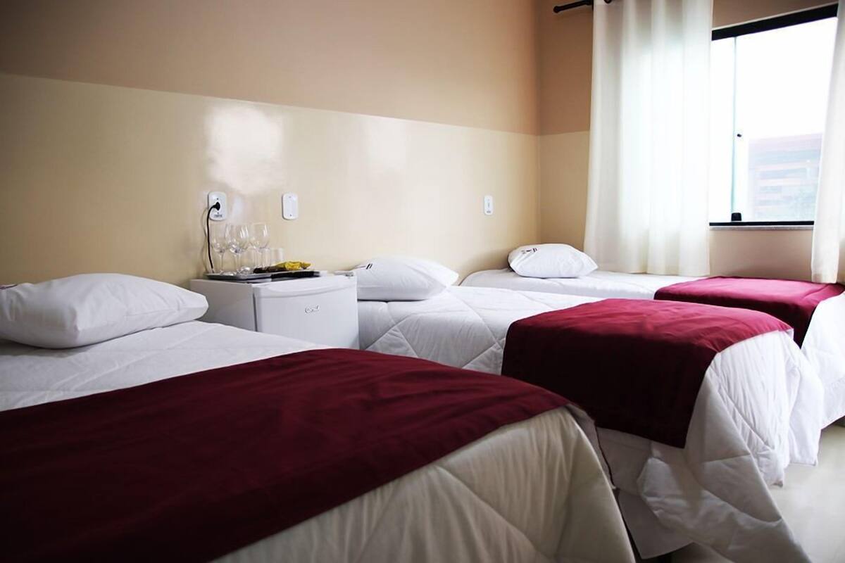 Een hotelkamer met twee bedden, elk voorzien van witte lakens en rode plaids, een nachtkastje met een lamp, en een raam met witte gordijnen.