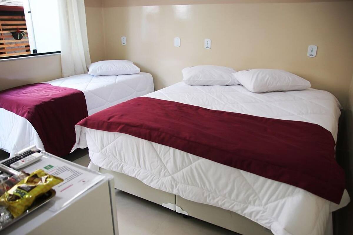 Een hotelkamer met twee eenpersoonsbedden, één met een rode deken en de ander met een witte deken. Er staat een klein tafeltje met een afstandsbediening en een zakje snacks.