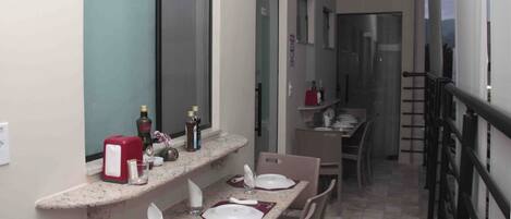 Un pasillo estrecho con una zona de cocina a la derecha, que incluye una mesa de comedor con platos y vasos, y una encimera con botellas y un electrodoméstico rojo.