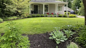 A two-story house with a covered porch, surrounded by a well-maintained lawn and various plants. - In-Town Saugatuck  “Magical Backyard” with Fire Pit Walk to Town (Saugatuck)