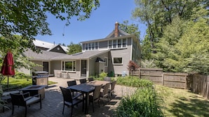 A two-story house with a patio area featuring a grill, outdoor seating, and a wooden fence. - In-Town Saugatuck  “Magical Backyard” with Fire Pit Walk to Town (Saugatuck)