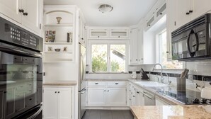 A modern kitchen with white cabinetry, a granite countertop, stainless steel appliances, and a window providing natural light. - In-Town Saugatuck  “Magical Backyard” with Fire Pit Walk to Town (Saugatuck)