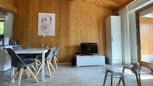 A modern living room with a wooden ceiling, a dining area with a table and chairs, a television on a white stand, and a glass table with a decorative item.
