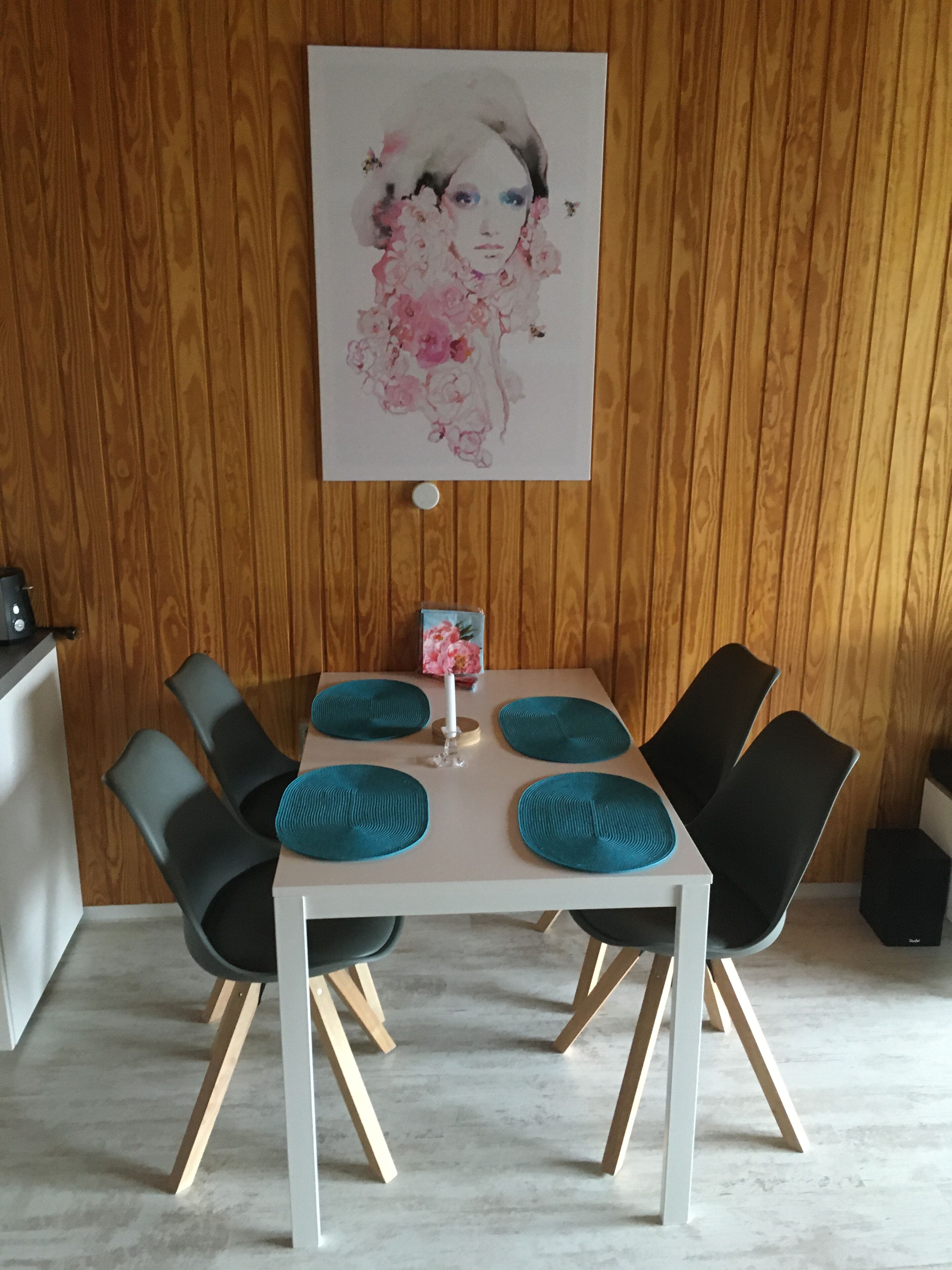 Een eethoek met een witte tafel, vier zwarte stoelen, blauwgroene placemats en een kunstwerk in een lijst aan de muur.