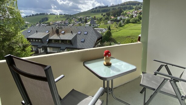 A balcony with a table and chairs, overlooking a town with houses and green hills.