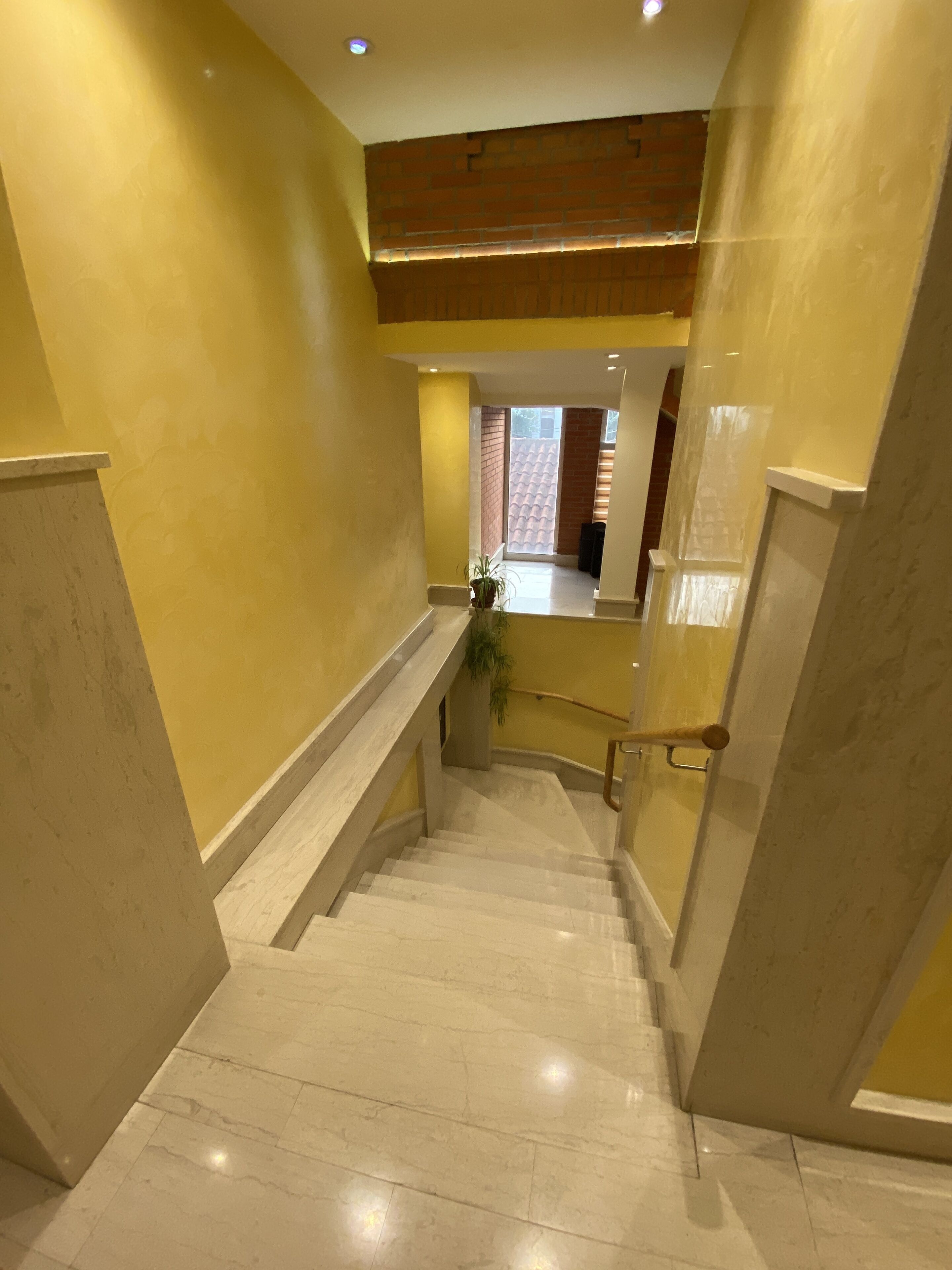 Una escalera con peldaños de mármol y paredes amarillas que conduce a una habitación con pared de ladrillo y una ventana.