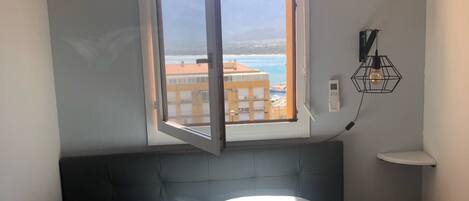 Un dormitorio con cama, ventana que ofrece vistas abiertas a edificios y una playa, una lámpara de pared y una pequeña mesa redonda.