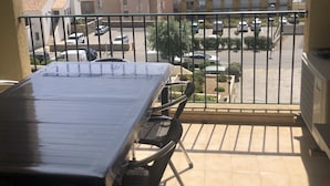 A balcony with a table and chairs, overlooking a parking area and buildings with a view of the sea and mountains.
