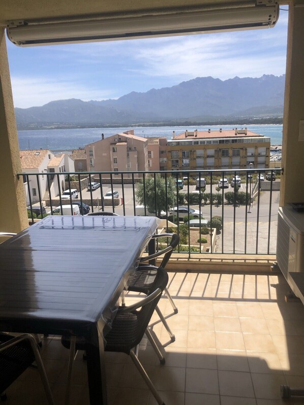 Uma varanda com mesa e cadeiras, com vista para um estacionamento e edifícios, onde se vê o mar e as montanhas.