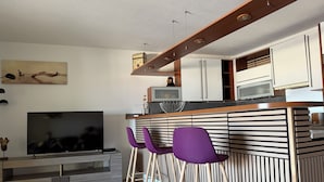 A modern kitchen with a wooden countertop, a built-in oven, and a microwave. There are two purple bar stools and a TV on a wooden stand.