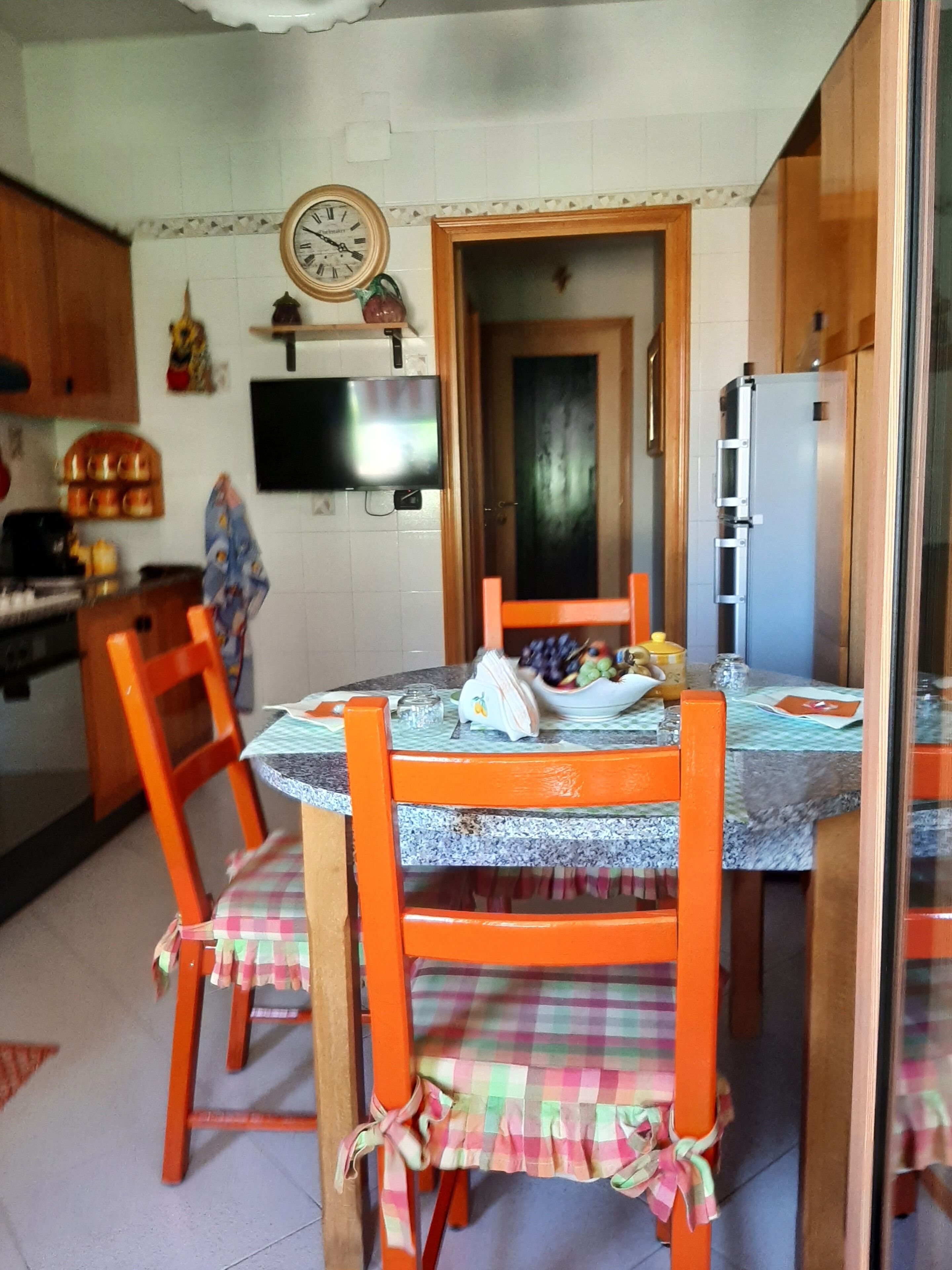 Een keuken met een eettafel, stoelen, een klok, een televisie en een koelkast.